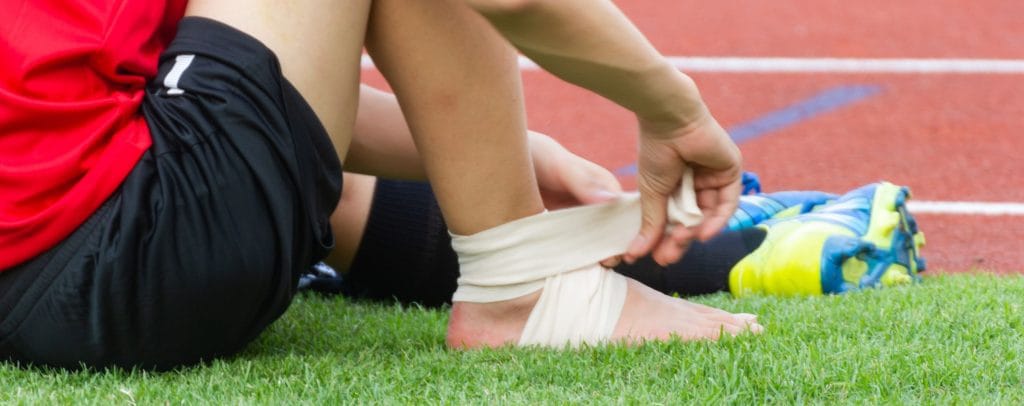 An athlete sitting on the grass field and wrapping their sports injury with bandages