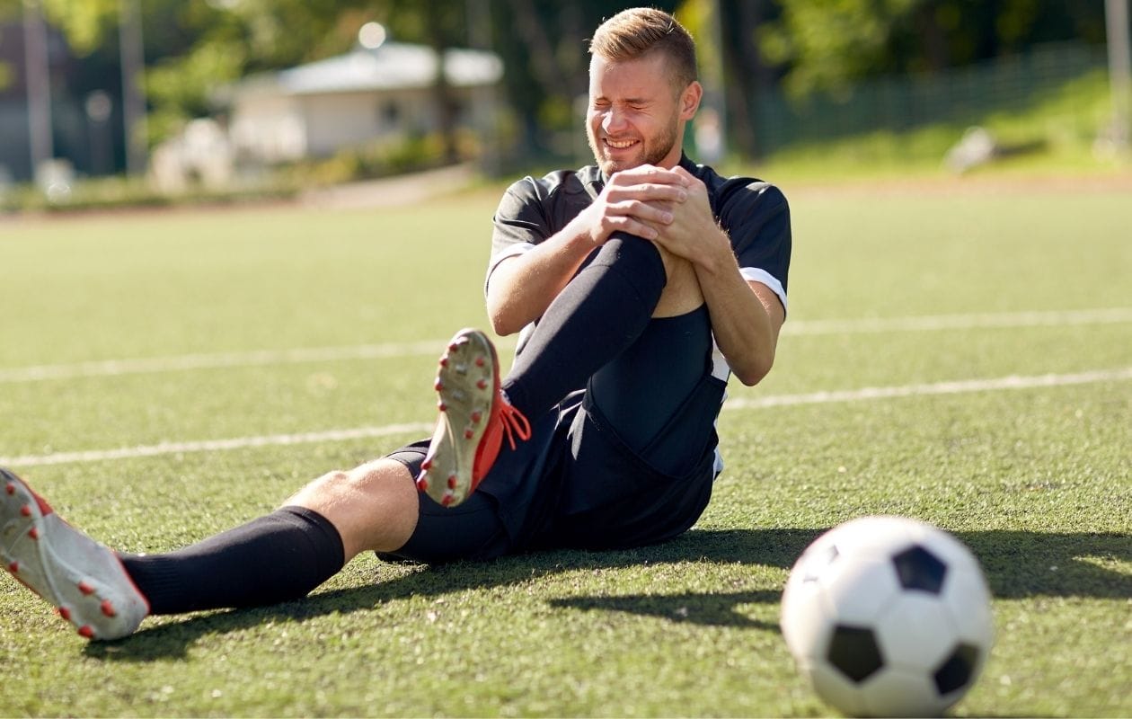 Male footballer sitting on a pitch in pain gripping his knee, indicating an ACL or knee ligament injury