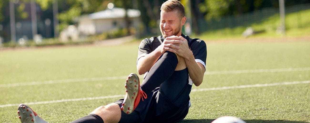 Male footballer sitting on a pitch in pain gripping his knee, indicating an ACL or knee ligament injury