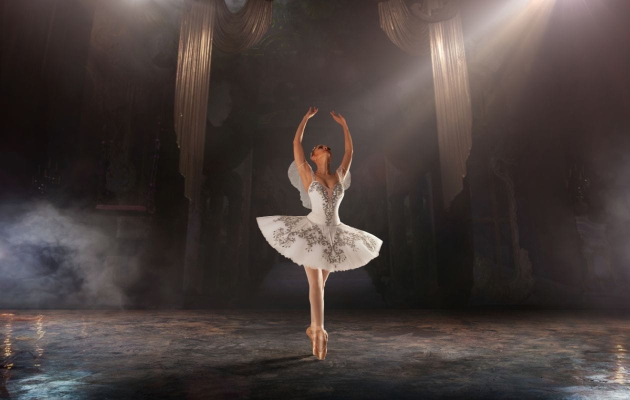 Ballet dancer in a white tutu posed with both arms raised on stage, demonstrating a confident return to performance after hypermobility physiotherapy
