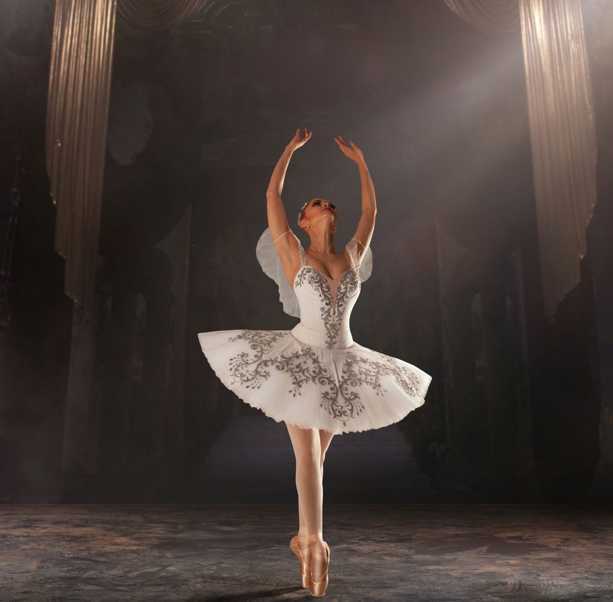 Ballet dancer in a white tutu posed with both arms raised on stage, demonstrating a confident return to performance after hypermobility physiotherapy