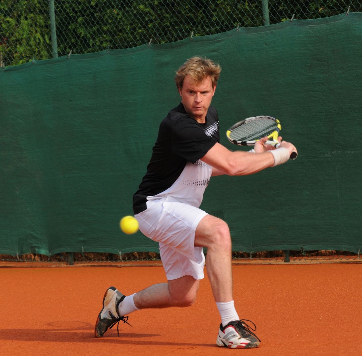 Male tennis player on a clay court hitting a two-handed backhand, demonstrating the repetitive shoulder loading common in racket sports