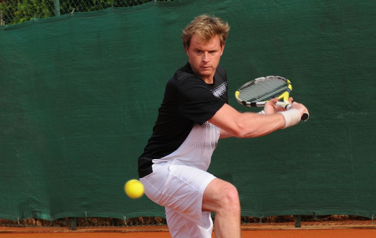 Male tennis player on a clay court hitting a two-handed backhand, demonstrating the repetitive shoulder loading common in racket sports