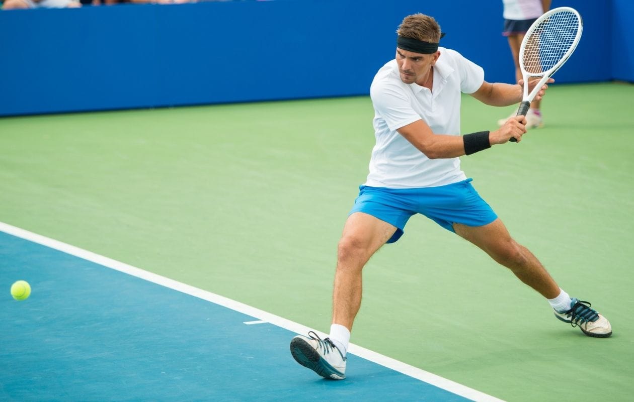 Male tennis player on a court hitting a low backhand, demonstrating the rotational and bending loads linked to sports spine injuries