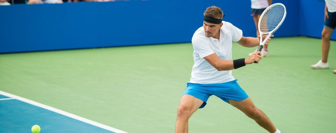 Male tennis player on a court hitting a low backhand, demonstrating the rotational and bending loads linked to sports spine injuries